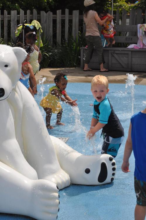 Children play in a splash pad, with a large polar bear sculpture and water fountains in a sunny outdoor setting.