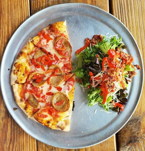 A slice of pepperoni pizza with red peppers, served alongside a fresh salad with bacon and shredded cheese on a metal plate.
