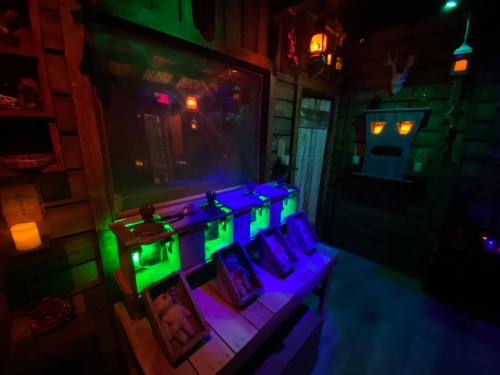 A dimly lit room with eerie decorations, featuring glowing lights, a large mirror, and several shadowy figures in display cases.