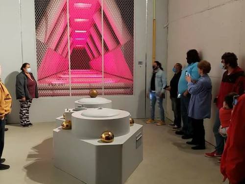 A group of people in a gallery, observing an installation with pink lighting and golden spheres on white pedestals.