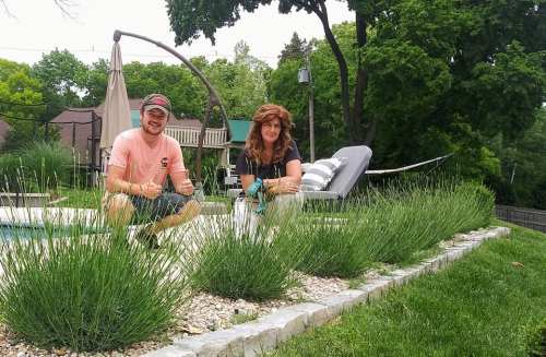 Two people sit on a stone border by a pool, surrounded by greenery and lavender plants, smiling at the camera.