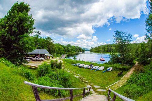 A serene lakeside scene with boats, a dock, and lush greenery under a partly cloudy sky.