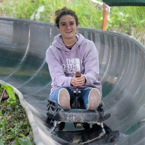 A young woman in a purple hoodie smiles while riding a luge on a winding track surrounded by greenery.