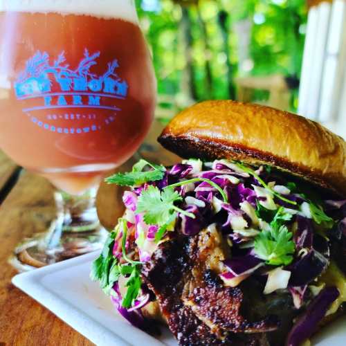 A close-up of a pulled pork sandwich topped with slaw and cilantro, alongside a glass of pink beverage on a wooden table.