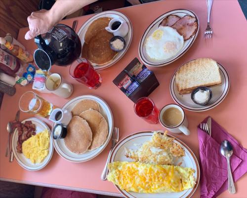 A breakfast spread featuring pancakes, eggs, bacon, toast, and drinks on a pink table.