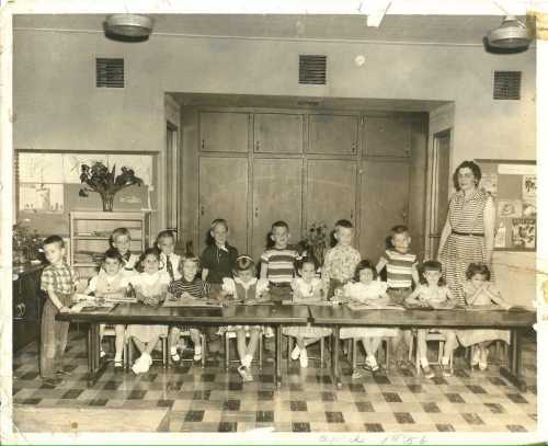 A black-and-white photo of a classroom with children at a long table and a teacher standing beside them.