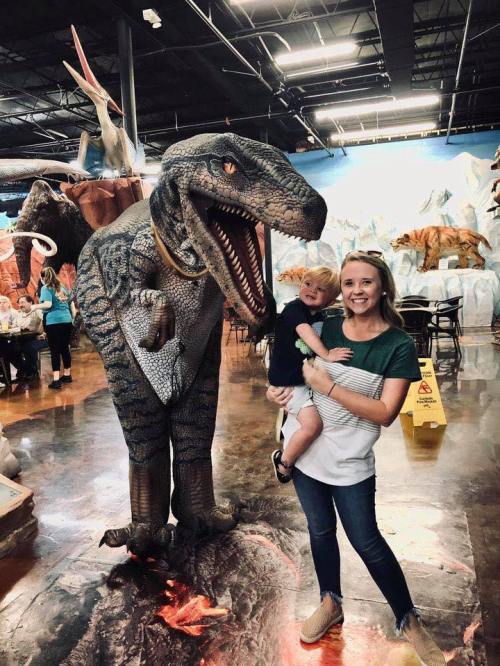 A woman holds a child next to a large dinosaur statue in a colorful indoor setting.
