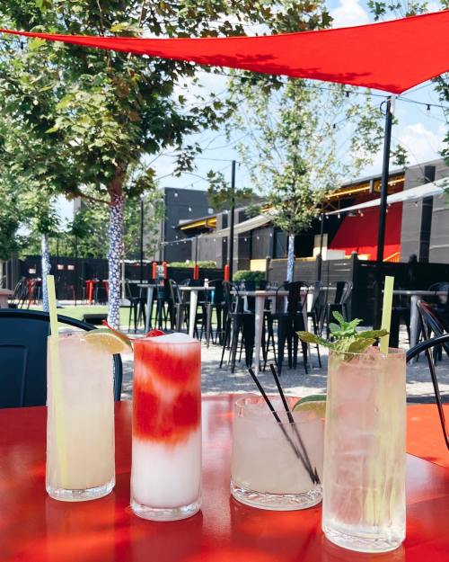 Four colorful cocktails on a red table, with a sunny outdoor setting and trees in the background.
