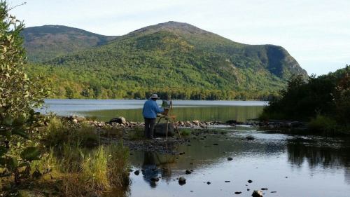 A painter stands by a calm lake, surrounded by mountains and lush greenery, capturing the serene landscape on canvas.