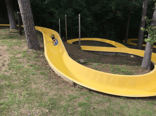 A child slides down a yellow water slide winding through a wooded area, surrounded by trees and grass.