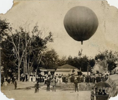 A historic scene of a crowd watching a hot air balloon ascending, with trees and buildings in the background.