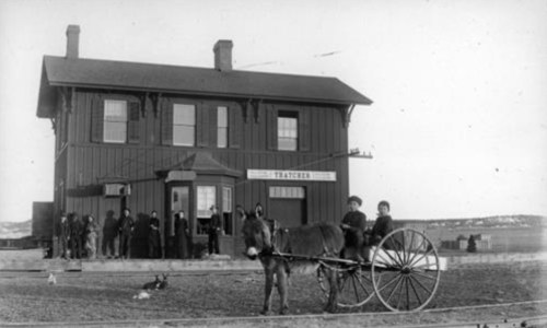 Historic black-and-white photo of a train station with a horse-drawn cart and people gathered outside.