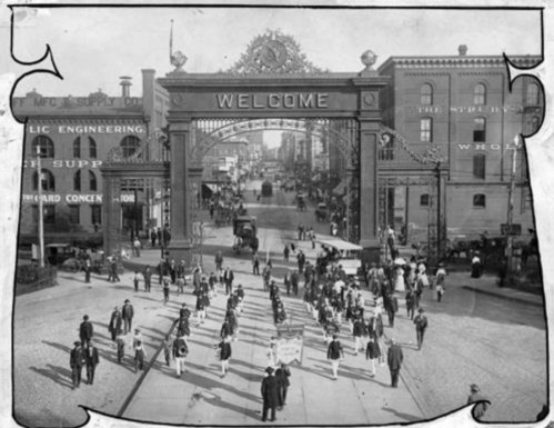 Historic black-and-white photo of a bustling street scene with a large "WELCOME" arch and crowds of people.