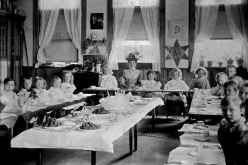 A vintage classroom scene with children seated at tables, enjoying a meal, and a teacher supervising.