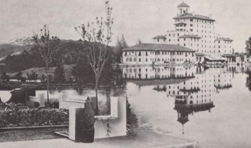 Historic black-and-white photo of a large hotel by a lake, surrounded by trees and mountains. Reflections in the water.