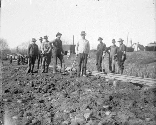 A group of men in hats and work clothes stand on a dirt road, holding tools, with buildings and vehicles in the background.