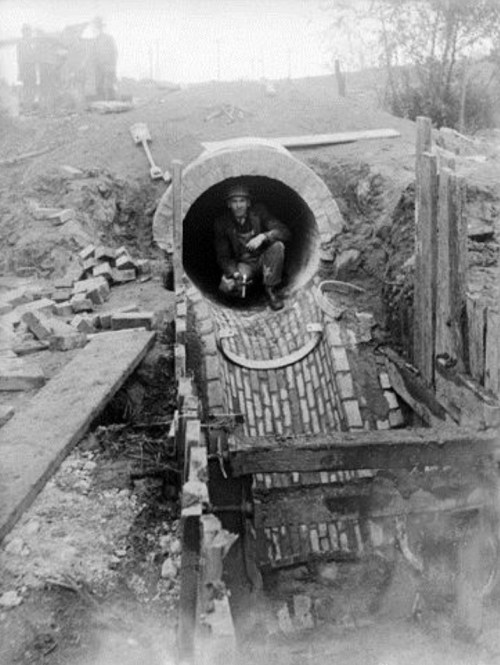A worker sits inside a large concrete pipe at a construction site, surrounded by bricks and earth.