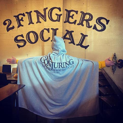 A person in a gray hooded cape stands with arms outstretched in front of a wall that reads "2 FINGERS SOCIAL."