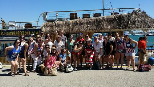 A large group of people in festive attire poses in front of a boat with a thatched roof on a sunny day.
