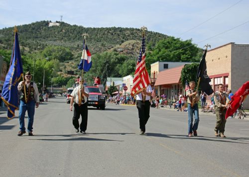 A parade with flag bearers walking down a street, surrounded by spectators and buildings, set against a hilly backdrop.