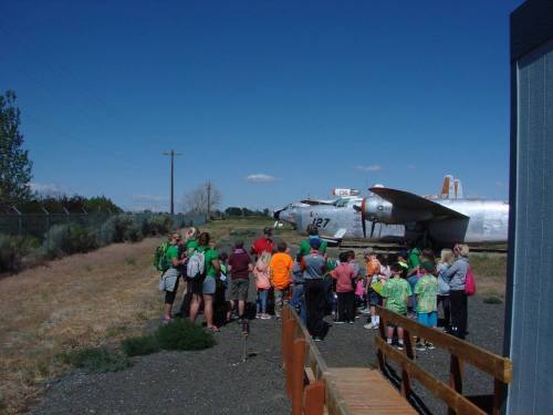 A group of children gathers near an old airplane on a sunny day, with trees and a fence in the background.