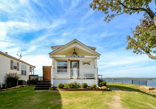 A charming lakeside cottage with a porch, surrounded by greenery and a scenic view of the water under a blue sky.