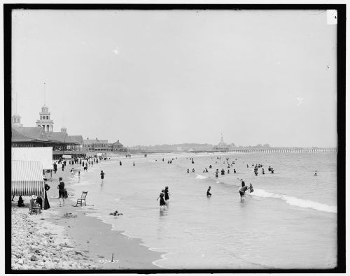 A vintage beach scene with people swimming and playing in the water, alongside a boardwalk and pier in the background.