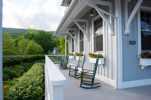 A charming gray house with rocking chairs on the porch, surrounded by greenery and flower boxes.