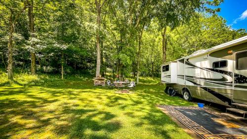 A sunny campsite with an RV parked on green grass, surrounded by trees and a picnic area with a table and chairs.