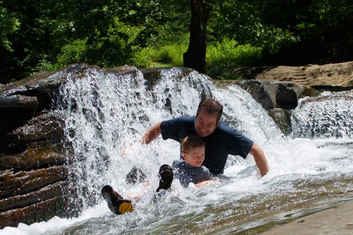 A man and a boy play in a waterfall, splashing water and enjoying a sunny day outdoors.