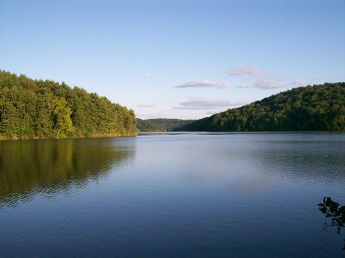A serene lake surrounded by lush green hills under a clear blue sky with a few clouds.