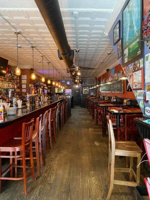 A cozy bar interior with wooden floors, a long counter, and dim lighting, featuring stools and tables along the walls.