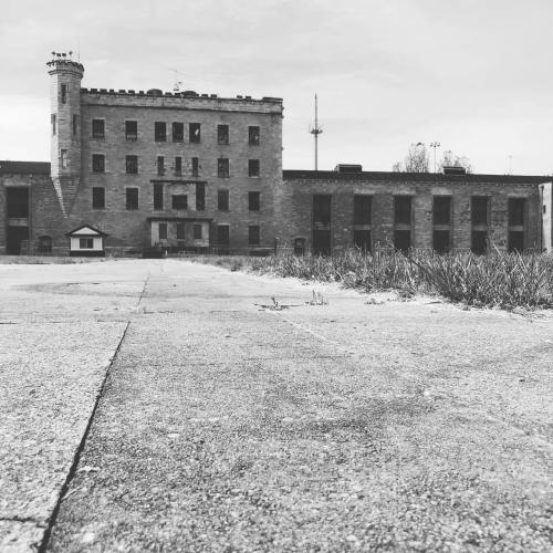 Abandoned stone building with a tower, surrounded by overgrown grass and a cracked pavement in black and white.