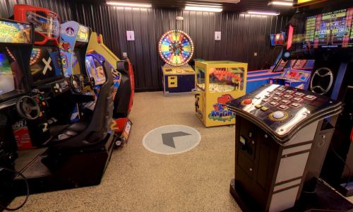 A colorful arcade room featuring various games, including racing and a prize wheel, with a circular directional marker on the floor.