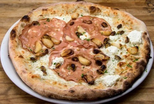 A pizza topped with slices of meat, roasted garlic, ricotta, and pistachios on a golden crust.