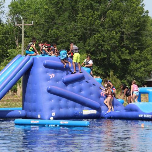 A group of children climbing and playing on a large blue inflatable water slide and climbing structure in a lake.