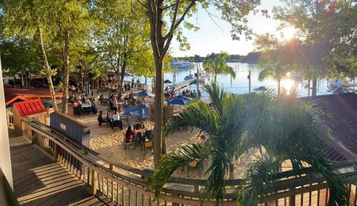 A scenic view of a waterfront restaurant with outdoor seating, surrounded by trees and boats on the water at sunset.