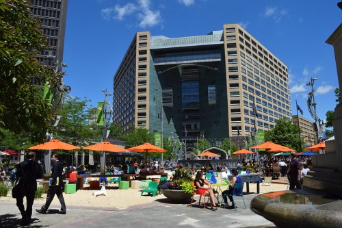 A bustling urban plaza with sandy areas, colorful umbrellas, and a modern building in the background under a clear blue sky.