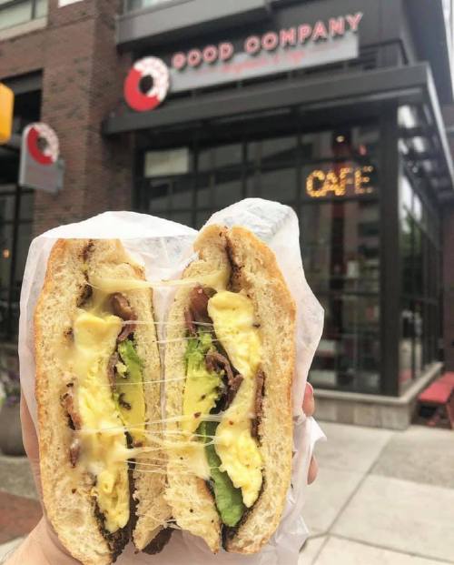 A hand holding a sliced sandwich with layers of egg, avocado, and meat, in front of a café.