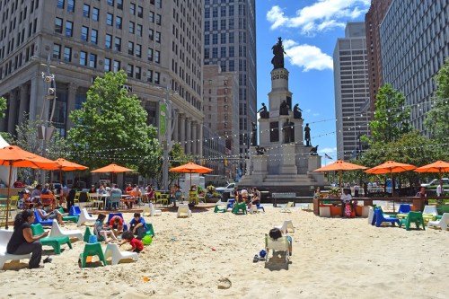 A sandy area with colorful umbrellas, people relaxing, and a monument in the background surrounded by tall buildings.