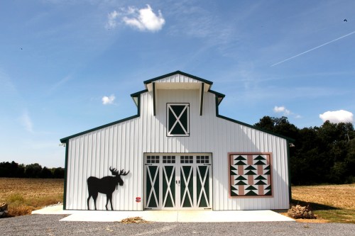 A white barn with green trim, featuring a moose silhouette and a geometric quilt pattern on the side, against a blue sky.