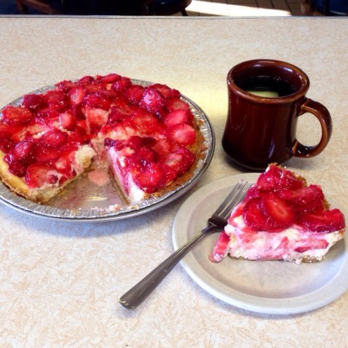A slice of strawberry pie on a plate next to a whole pie and a cup of coffee on a table.