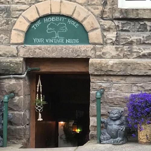 A stone entrance labeled "The Hobbit Hole" with vintage decor, featuring a frog statue and purple flowers nearby.