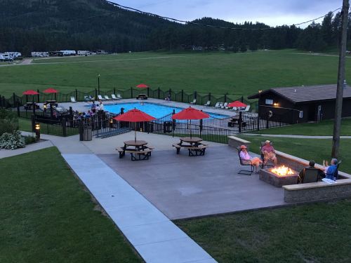 A scenic view of a pool area with red umbrellas, picnic tables, and a fire pit, surrounded by green hills and trees.
