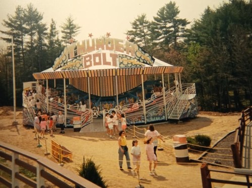A vintage amusement ride called "Thunder Bolt" with colorful signage, surrounded by trees and visitors in summer attire.