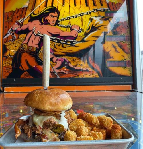 A delicious burger with cheese and jalapeños on a tray, with tater tots, set against a colorful retro arcade backdrop.