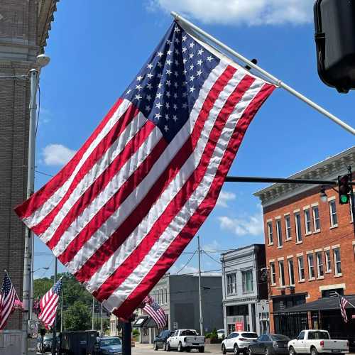 A large American flag waves in the breeze against a clear blue sky, with buildings and more flags in the background.