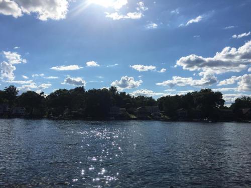 A serene lake scene with sparkling water, lush trees, and a bright blue sky dotted with fluffy clouds.