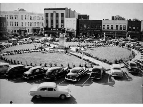 Historic black-and-white photo of a town square with cars parked around a central monument and surrounding buildings.