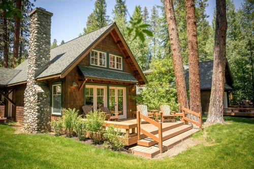 A cozy wooden cabin with a stone chimney, surrounded by tall trees and a green lawn, featuring a deck with chairs.